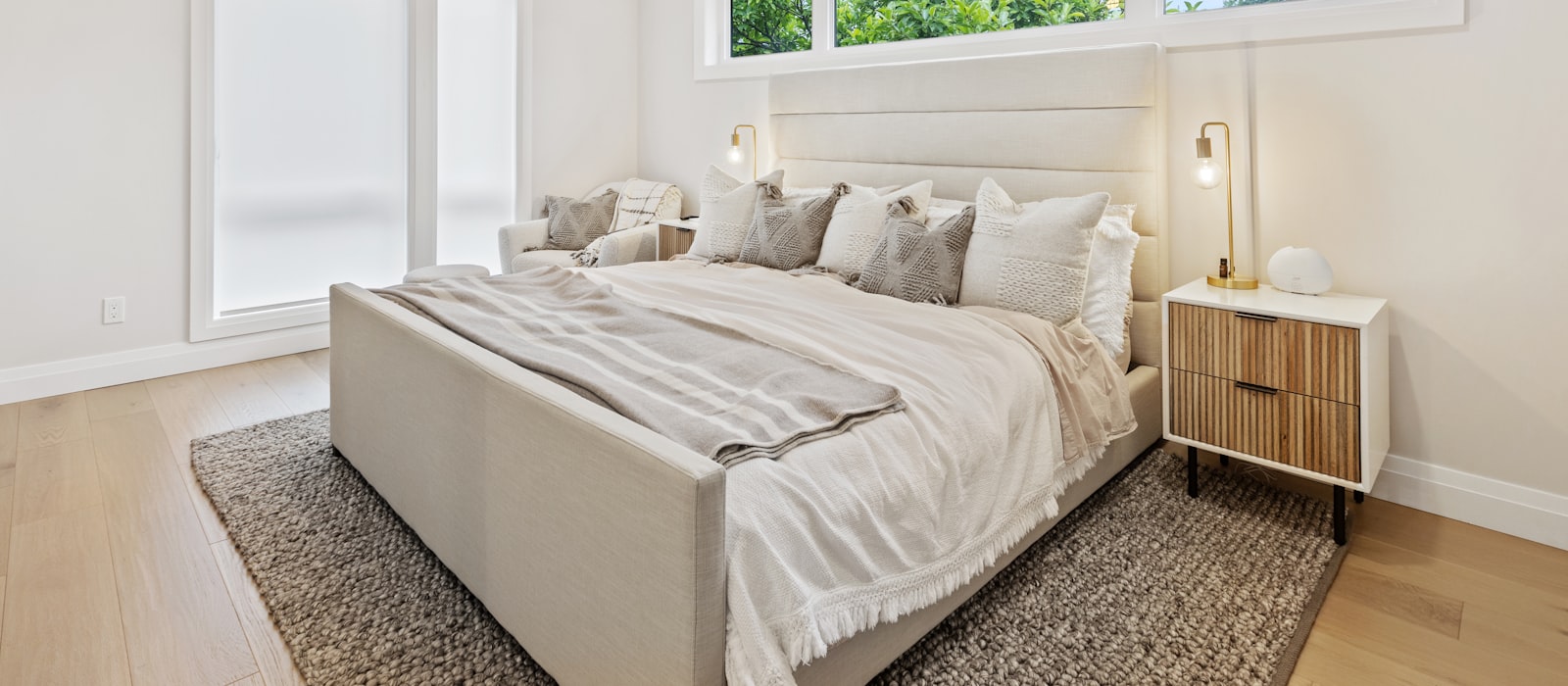 A neatly made bed with white linen and grey cushions in a bright bedroom, daylight from a window falling across a wood floor and rug