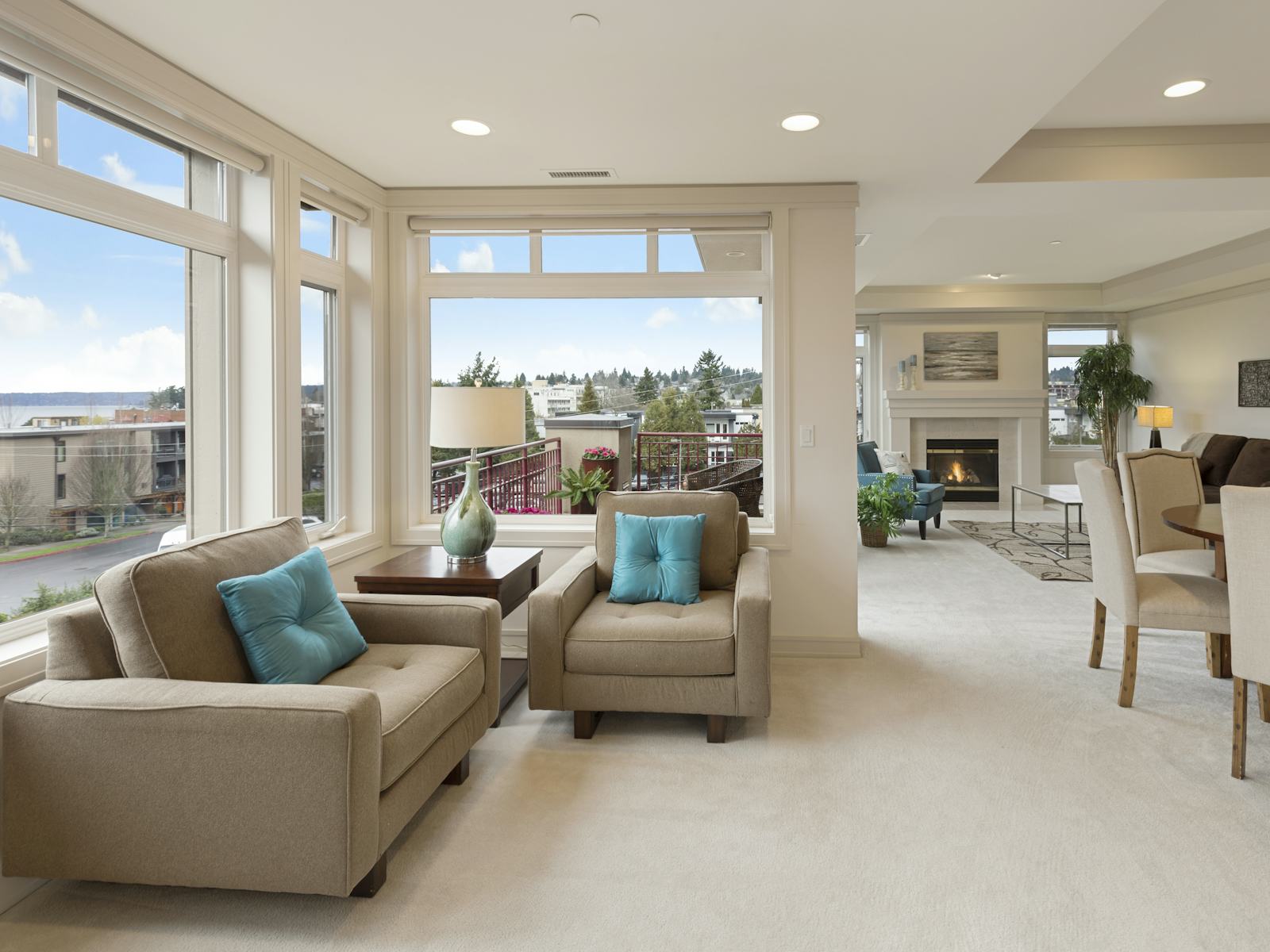 A bright, tidy living room with beige armchairs, turquoise cushions and large windows looking onto a balcony