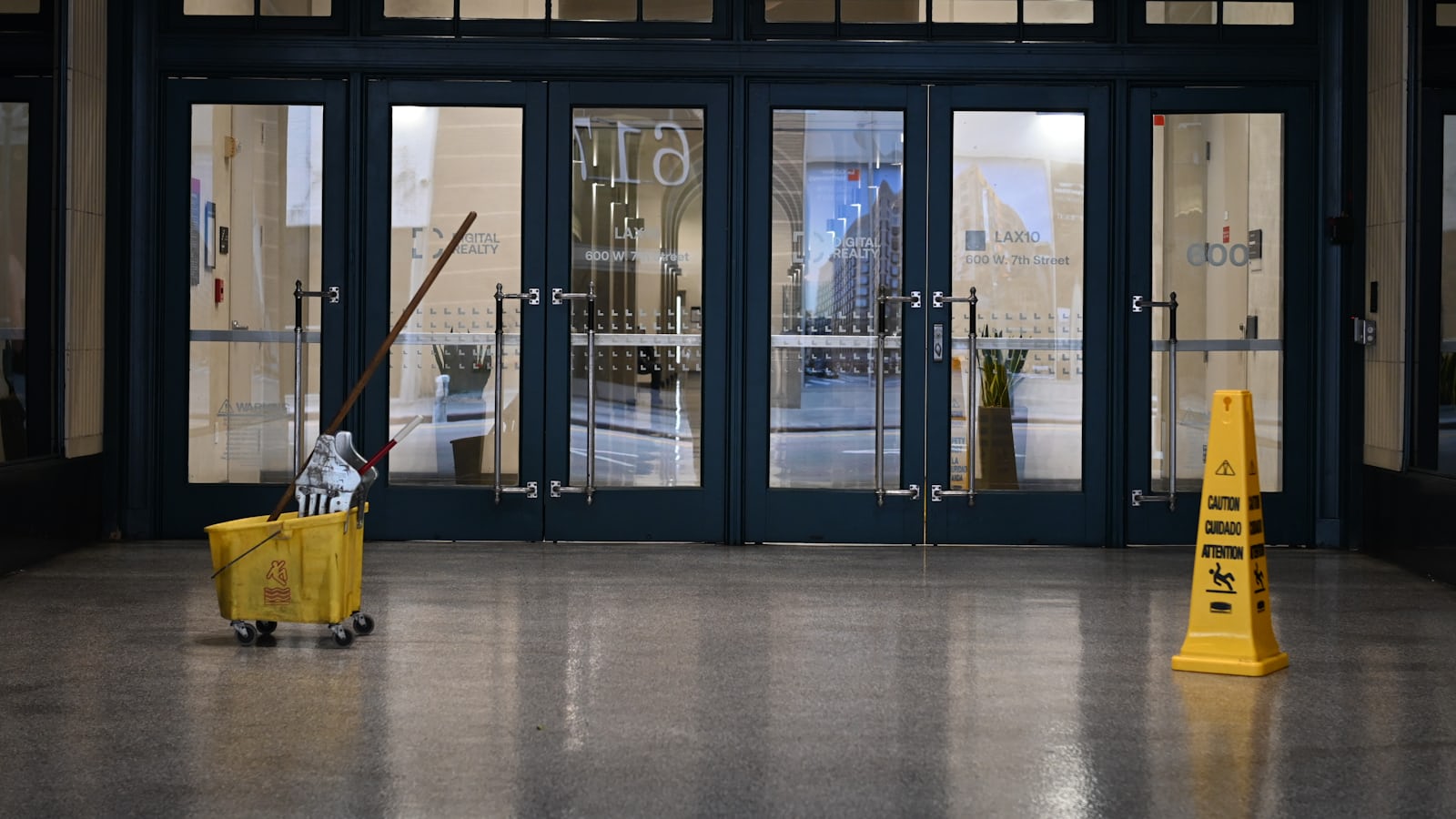 A yellow mop bucket with wringer and a yellow caution cone standing on a polished floor inside a glass building entrance